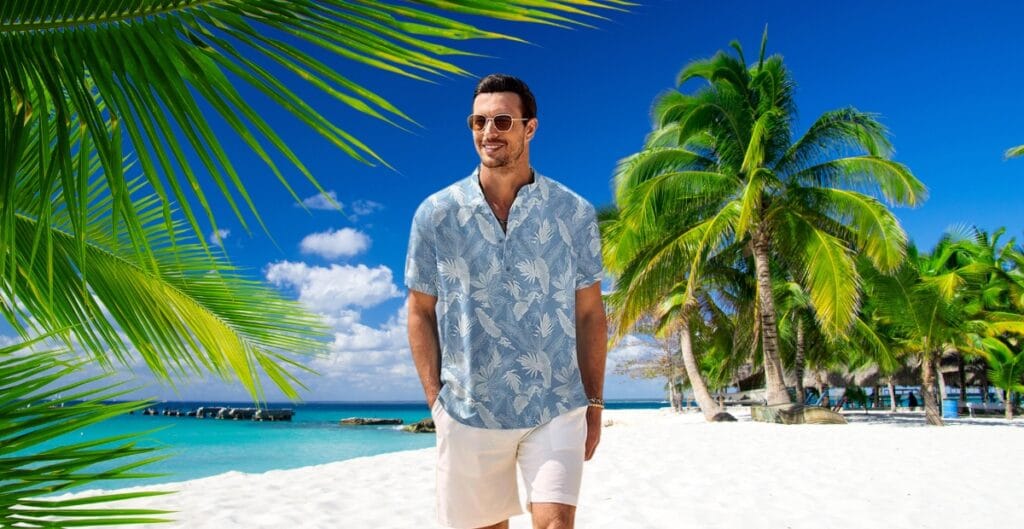 A man walking along a tropical beach with white sand, turquoise water, and lush green palm trees, wearing a light blue Hawaiian shirt with white floral patterns and white shorts