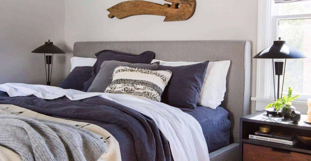 A neatly made bed with a light gray upholstered headboard, layered with dark gray, white, and patterned pillows, topped with a cozy gray blanket. On each side of the bed are black bedside lamps, and there is a window with white trim letting in natural light, along with a small black bedside table holding a plant and some books.