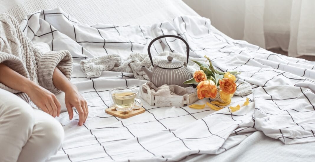 A cozy bed with a white and black checkered bedsheet, a person sitting at the edge, a teapot with a tray, a glass of tea or herbal infusion, and a small bouquet of orange flowers with fallen petals.