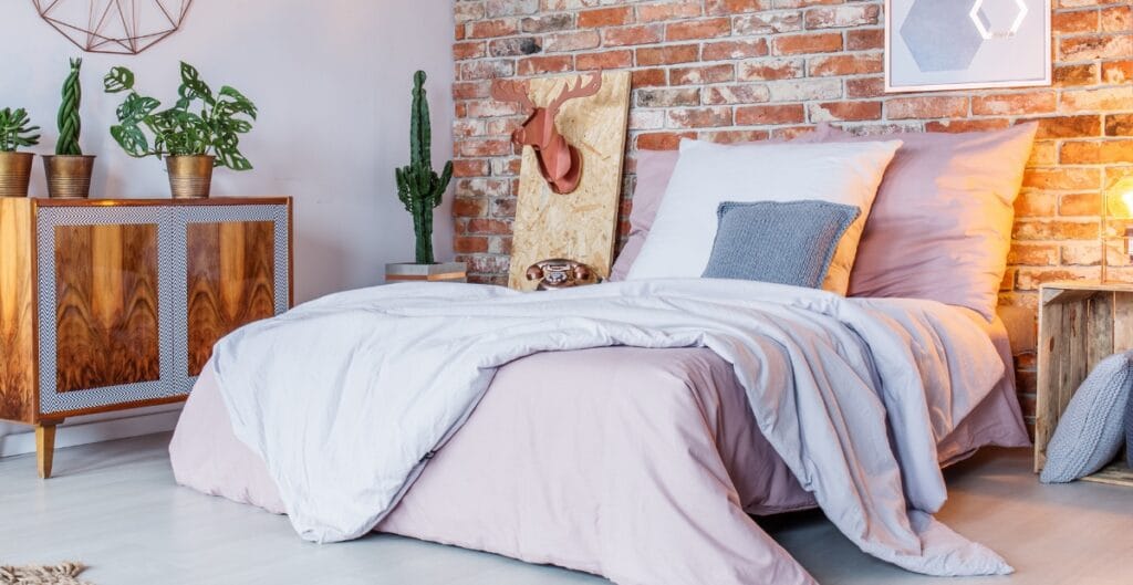 A cozy bedroom with a bed covered in light pink and white bedding, set against a brick wall with decorative elements, including a cactus plant, a wooden side table, and framed art.