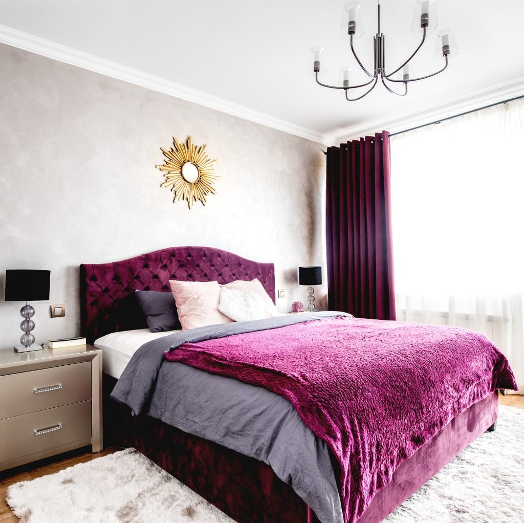 A stylish bedroom featuring a plush, deep maroon velour bed with an upholstered headboard, complemented by matching velour curtains, black bedside lamps, and a modern chandelier.