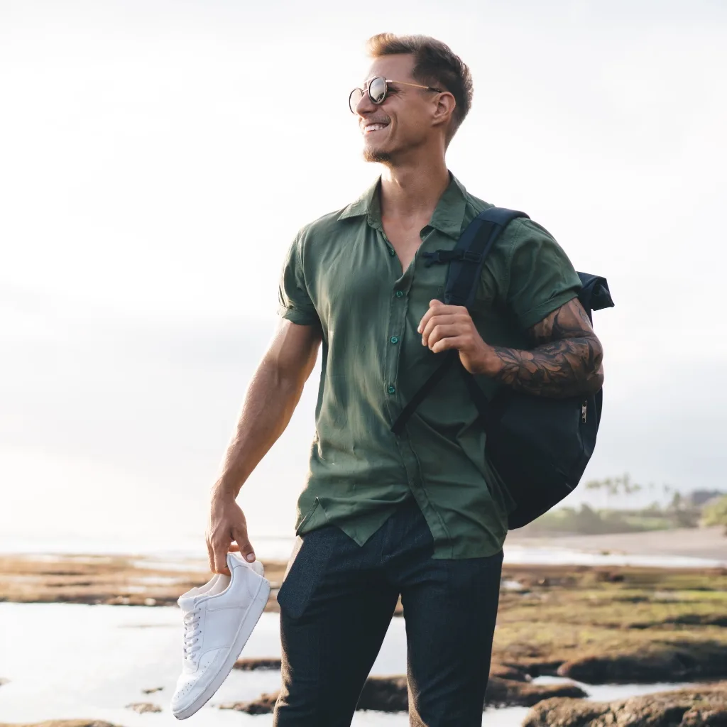 A man outdoors on a bright, sunny day, wearing a short-sleeved, lightweight, breathable summer shirt in green. He is smiling, carrying a backpack, and holding a pair of white sneakers.