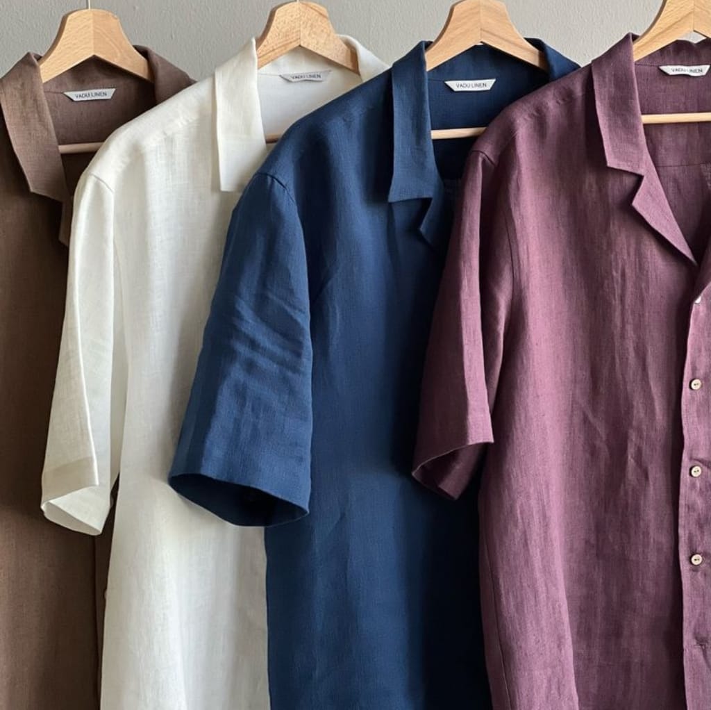 Four men's linen shirts in brown, white, navy blue, and maroon hanging on wooden hangers against a neutral background.