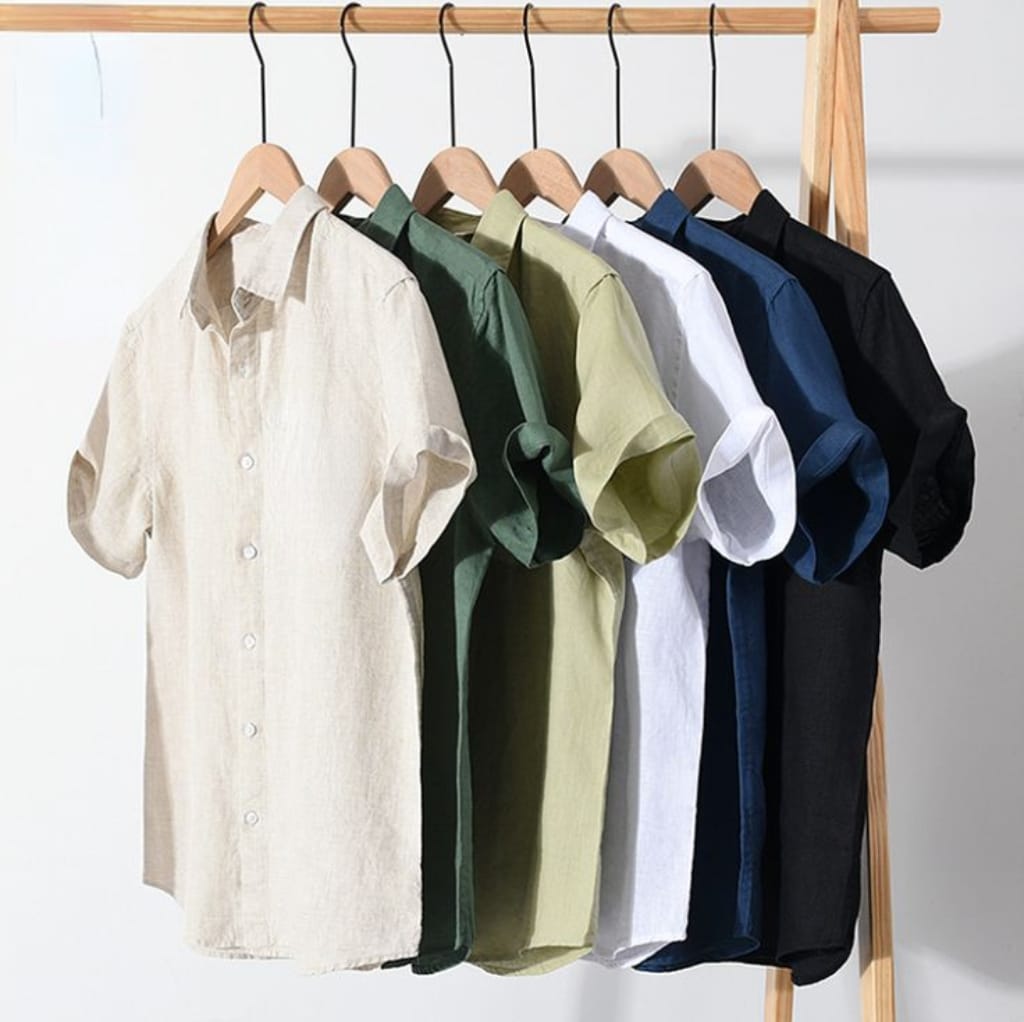 A row of six linen short sleeve shirts hanging on wooden hangers, displayed on a wooden rack. The shirts are in various summer-appropriate colors: cream, dark green, light green, white, navy blue, and black.