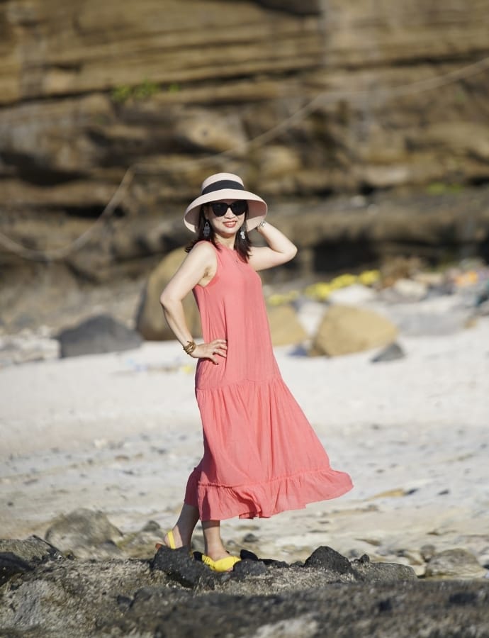 A woman in a coral tiered cotton dress, wearing a wide-brimmed hat and sunglasses, stands on a rocky beach with a sandy background and large rocks.