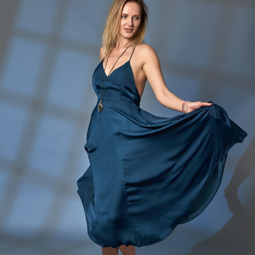 A woman wearing a flowing, deep blue satin maxi dress with thin spaghetti straps, holding out the skirt with one hand.