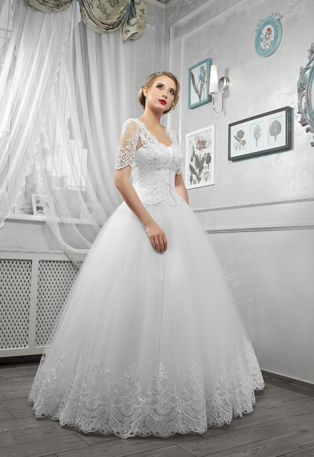 A woman wearing a classic ball gown wedding dress with lace details on the bodice and sleeves, standing indoors against a light-colored, elegantly decorated wall with framed artwork.