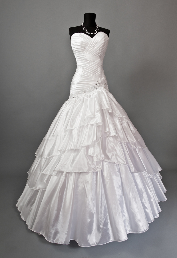 A sleek, elegant mermaid-style wedding gown displayed on a black mannequin. The dress features a fitted bodice with ruched detailing and a strapless sweetheart neckline, flowing into a voluminous layered skirt with satin ruffles.