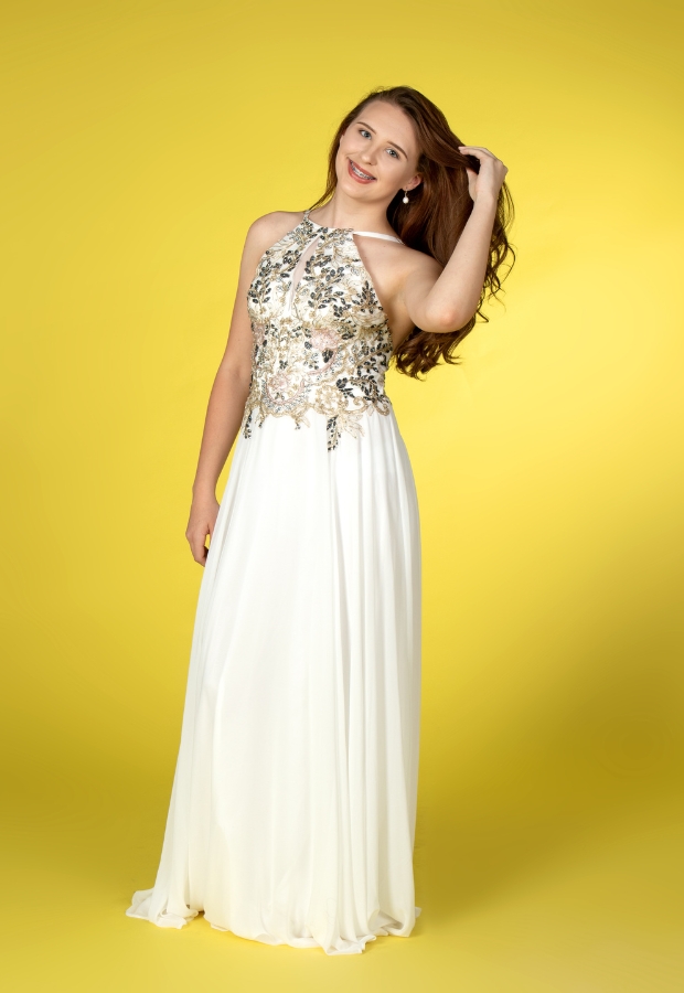 A young woman with long, wavy brown hair is wearing a two-piece prom dress against a bright yellow background. The top is sleeveless with intricate floral embroidery and beading, and the long, flowing skirt is white with soft pleats.