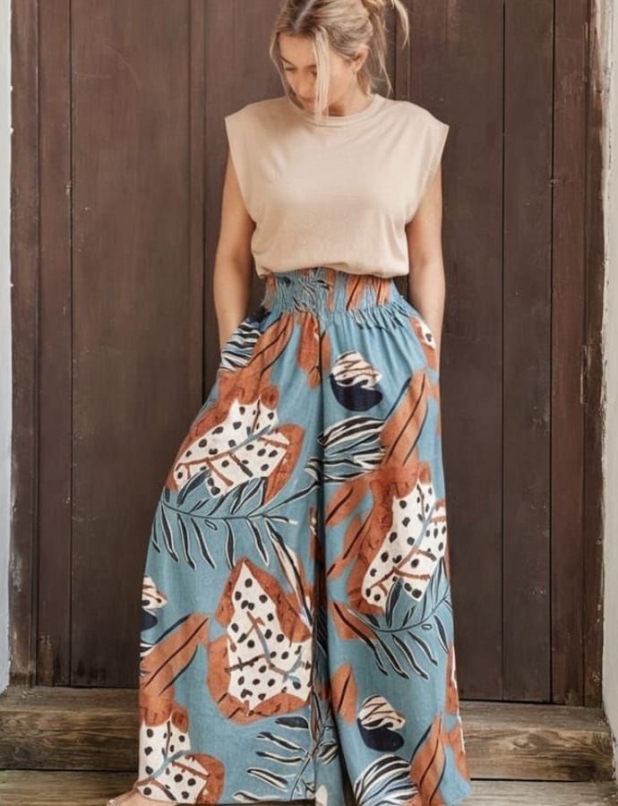 A woman standing in front of a wooden wall, wearing a sleeveless beige top tucked into high-waisted, wide-leg pants with a bold tropical and animal print pattern in shades of blue, white, black, and rust.