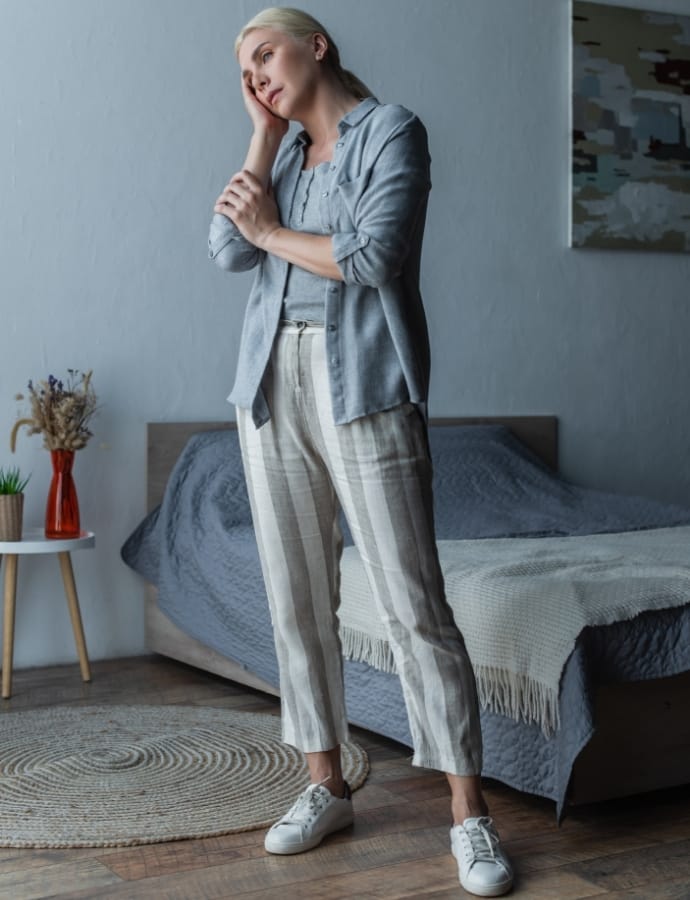 A woman with blonde hair is standing in a modern bedroom, wearing a light blue woven cotton shirt, striped plaid trousers, and white sneakers. She appears thoughtful with her hand resting on her face, and the room features a bed with a textured blanket, a round rug, and a small side table with dried flowers.