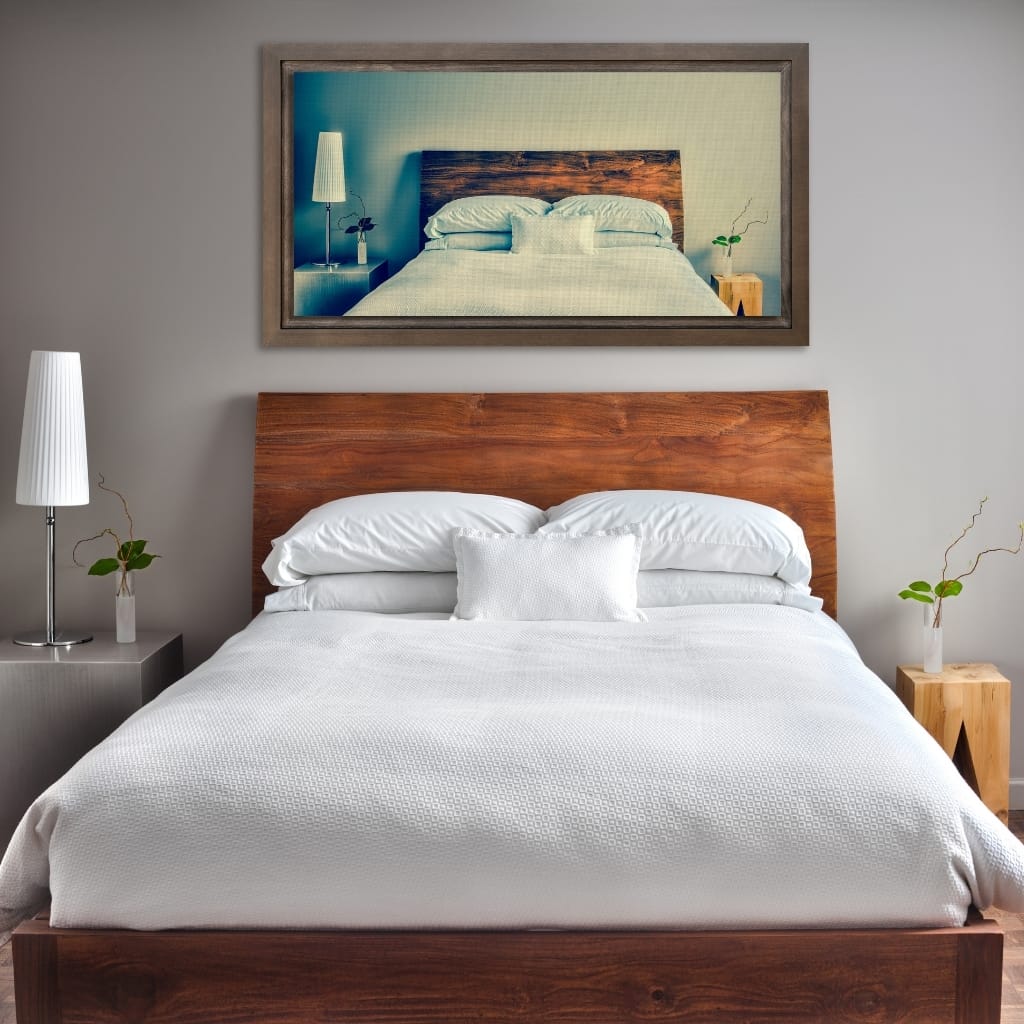 A modern bedroom with a large wooden bed frame and white bedding, flanked by two nightstands with minimalist lamps and small green plants. A framed picture of a bedroom scene hangs above the bed, set against a plain light gray wall.