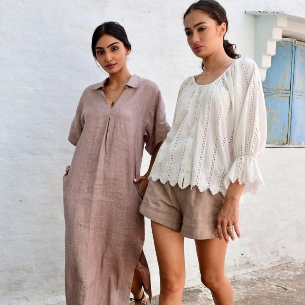 Two women standing against a white textured wall, one wearing a long, beige linen dress with a collar, and the other wearing a white, embroidered blouse with beige shorts.