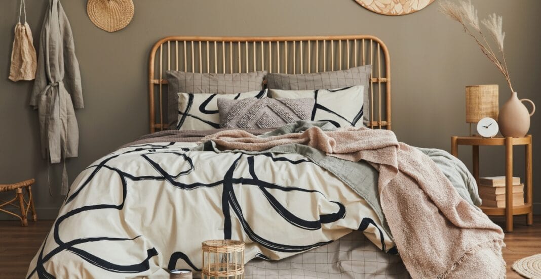 A neatly made bed with a rattan headboard, layered with modern patterned pillows, a sheet with bold black abstract lines, and a cozy pink blanket draped across. To the right, there is a wooden nightstand with a round clock, a white ceramic vase with dried branches, and a small lamp. The wall behind is painted in a neutral taupe color with a decorative round wall hanging.