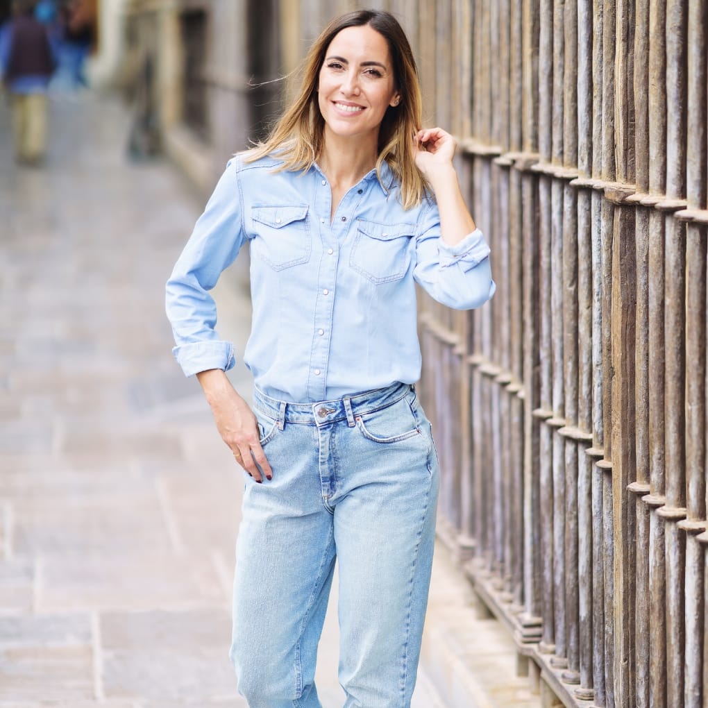 Woman in light blue chambray shirt and denim jeans standing outdoors near a wooden fence, smiling at the camera.