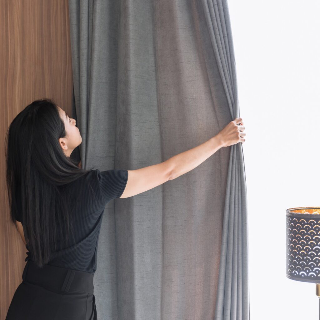 A woman with long dark hair, wearing a black shirt, is pulling aside a grey curtain to look outside in a well-lit room. A black decorative lamp with cut-out patterns is visible on the right side.
