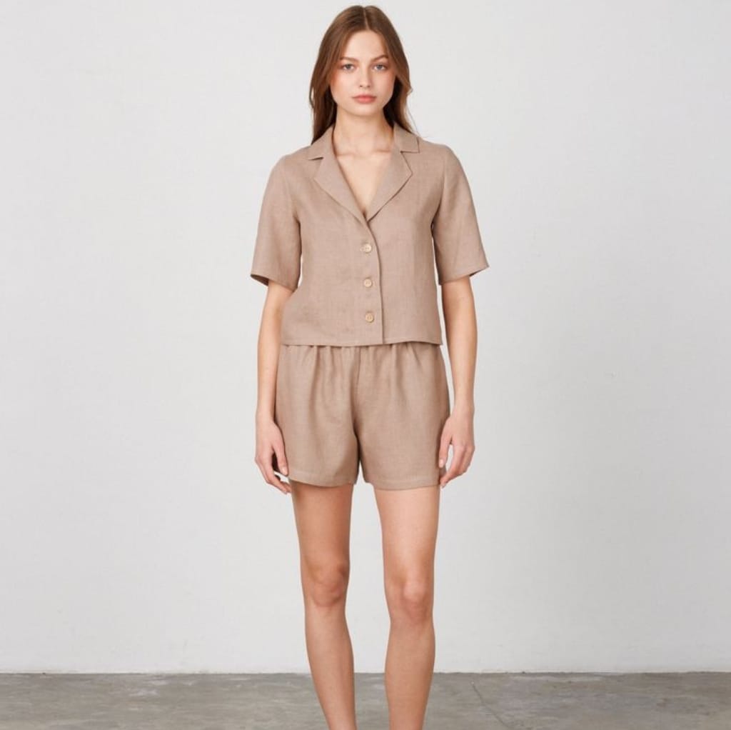 A woman wearing a beige linen short sleeve shirt and matching shorts, standing against a plain white background.