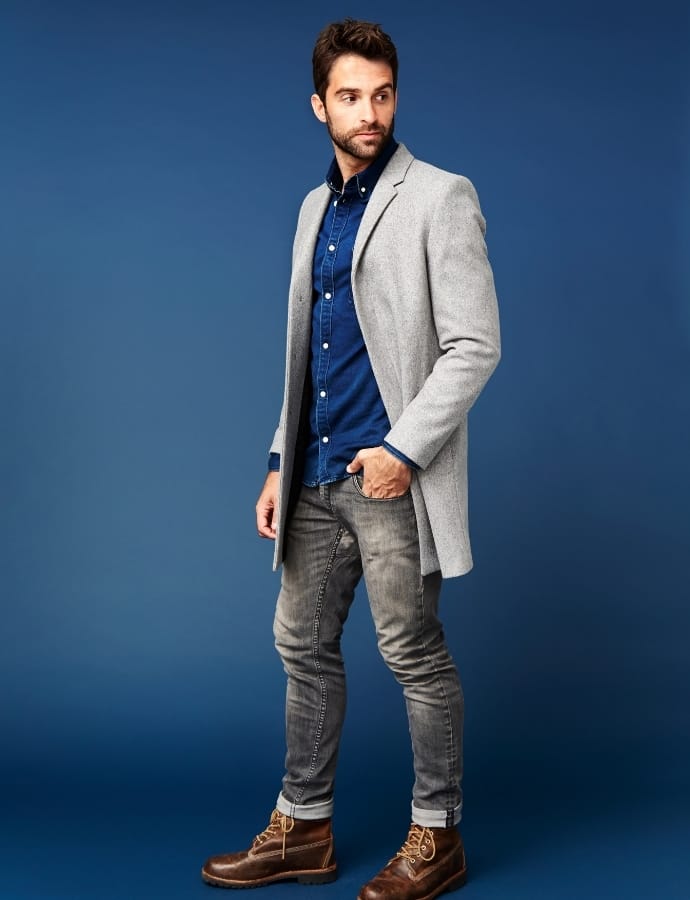 A man stands against a blue background wearing a light gray sport coat over a dark blue shirt, paired with gray jeans and brown boots.