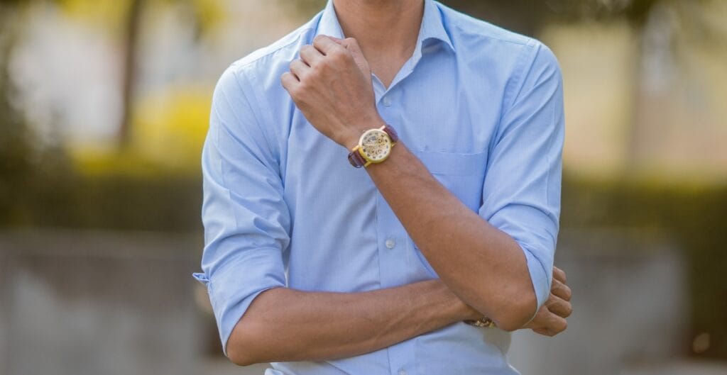 A person wearing a light blue chambray button-down shirt, with the sleeves rolled up, standing outdoors with trees in the background.