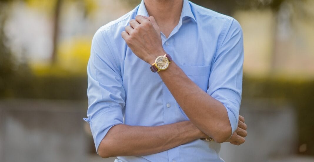 A person wearing a light blue chambray button-down shirt, with the sleeves rolled up, standing outdoors with trees in the background.