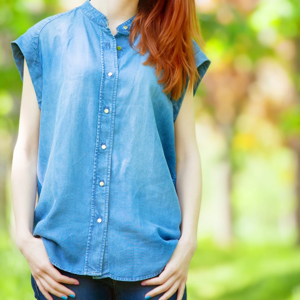 A person wearing a sleeveless chambray shirt with a stand collar, standing outdoors with a blurred green background.