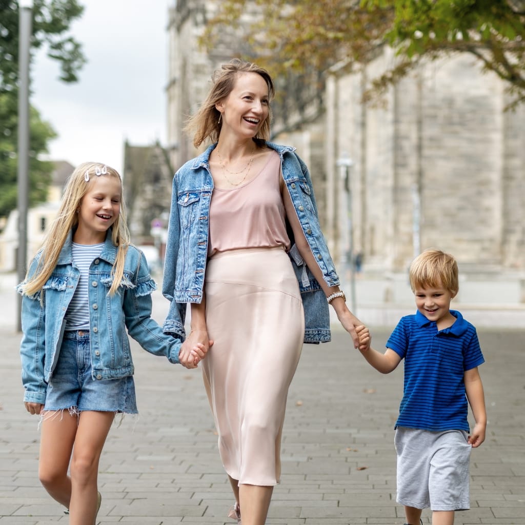 A smiling woman walking outdoors holding hands with a young girl and a young boy, all dressed in casual denim outfits, with trees and historic buildings in the background.