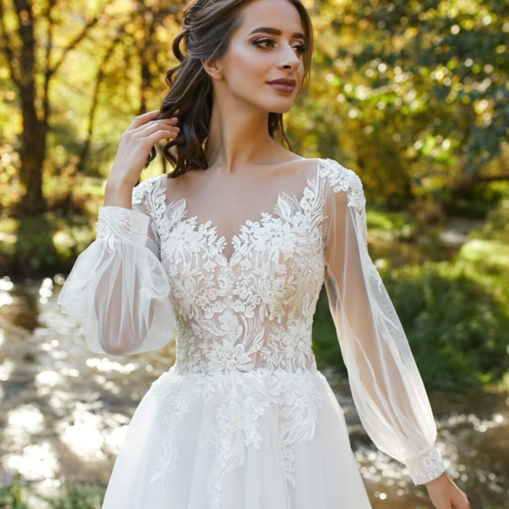 A woman wearing a beautiful wedding dress with intricate lace embroidery on the bodice and sheer, puffed long sleeves, standing outdoors with autumn foliage in the background.