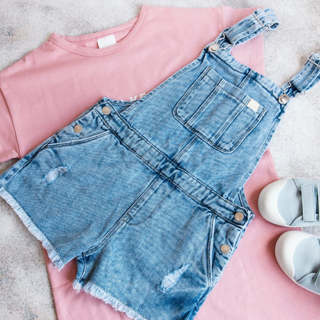 A pink t-shirt, a pair of denim bib overalls with distressed details, and white sneakers placed on a light textured surface.