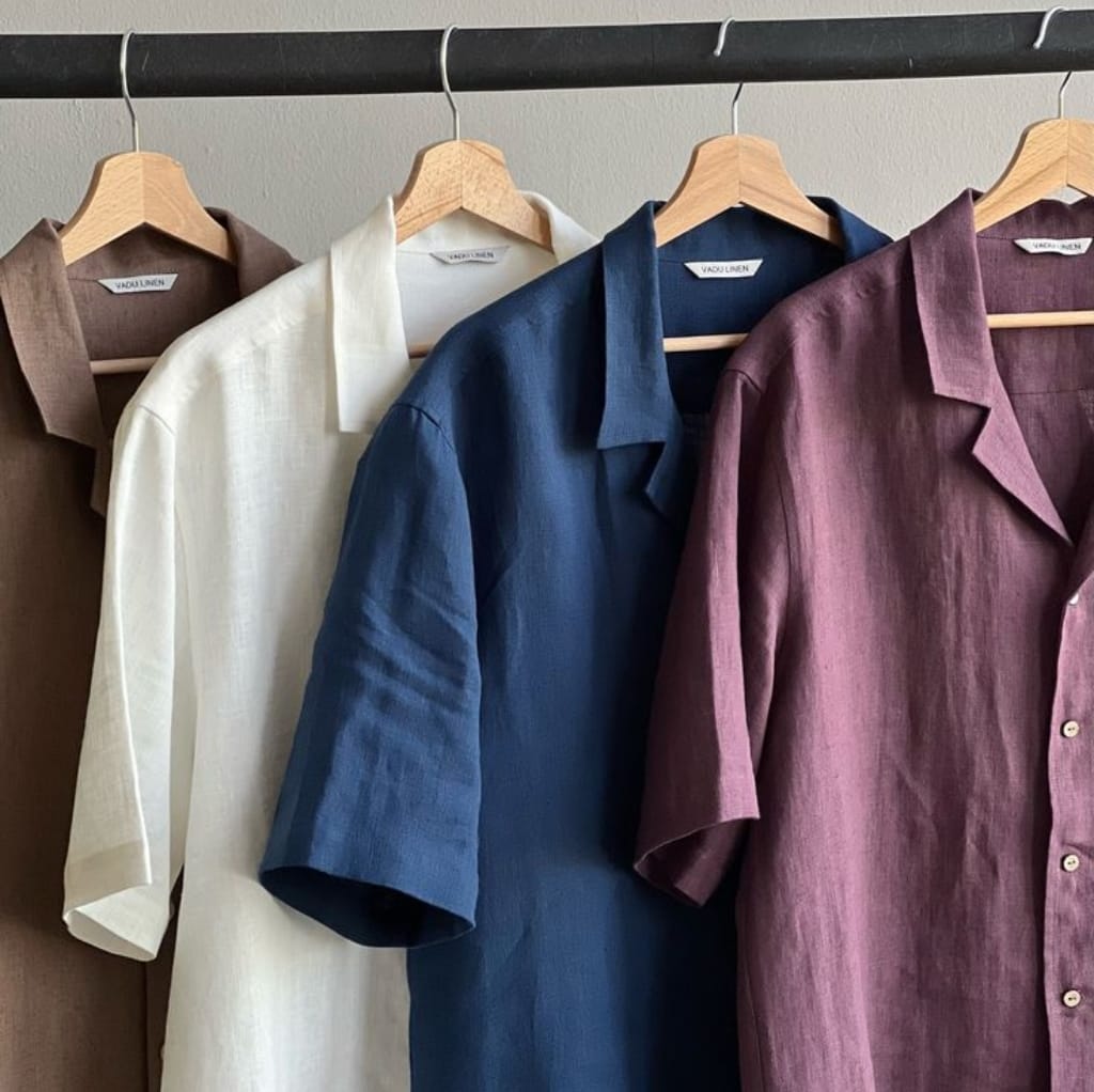 Four linen short-sleeve shirts in brown, white, blue, and mauve hanging on wooden hangers on a black clothing rack.