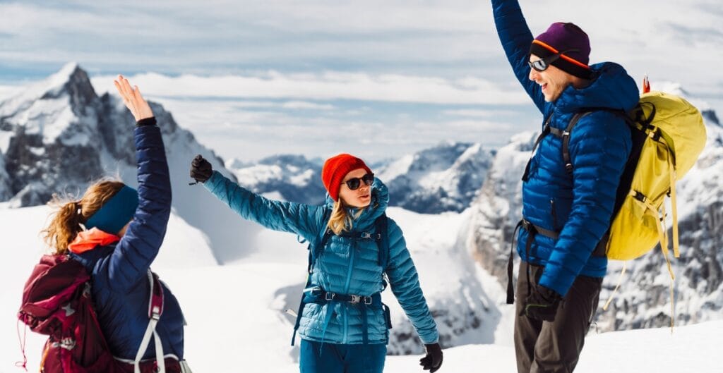 Three hikers in winter gear greeting each other on a snowy mountain landscape, with rugged peaks in the background