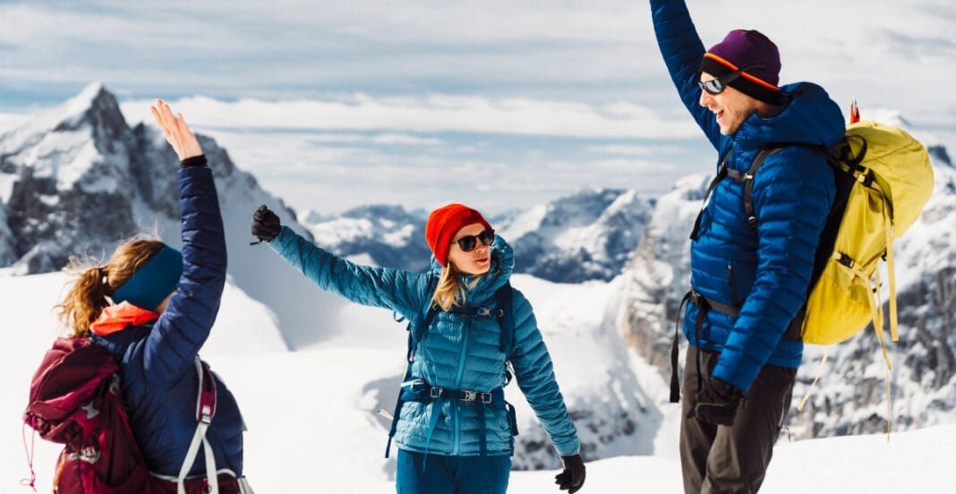 Three hikers in winter gear greeting each other on a snowy mountain landscape, with rugged peaks in the background