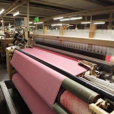 Close-up view of a textile manufacturing machine combing cotton fibers, with pink-striped fabric being processed on the rollers.