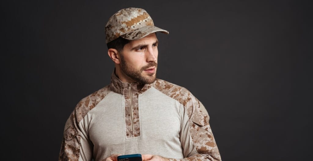 A man in military camouflage attire, holding a phone, looking to the side against a dark background.