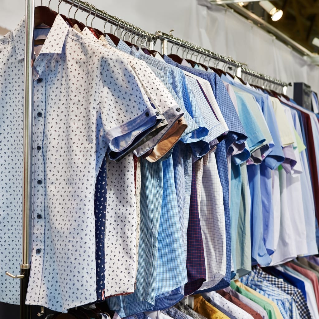 A row of men's linen shirts in various colors hanging on a clothing rack at a market or store, including shades of white, blue, light green, pastel, and patterned designs.