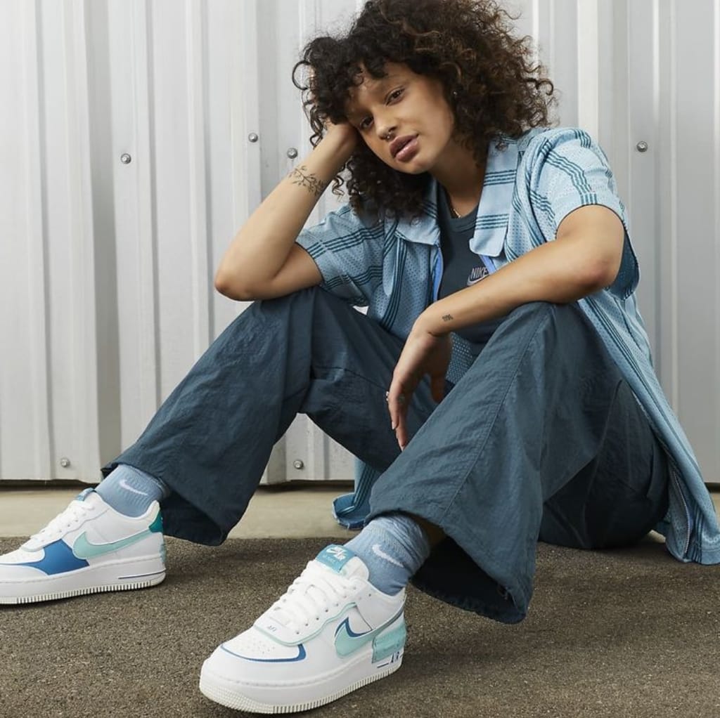 A young woman with curly hair, dressed in a gray and blue Nike outfit, is sitting on the ground against a metallic wall, wearing white Nike sneakers with pastel accents. Best Nike Sports Wear