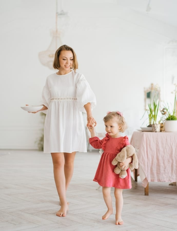 A woman in a white dress holding the hand of a young girl in a pink dress, walking barefoot in a bright, airy room.