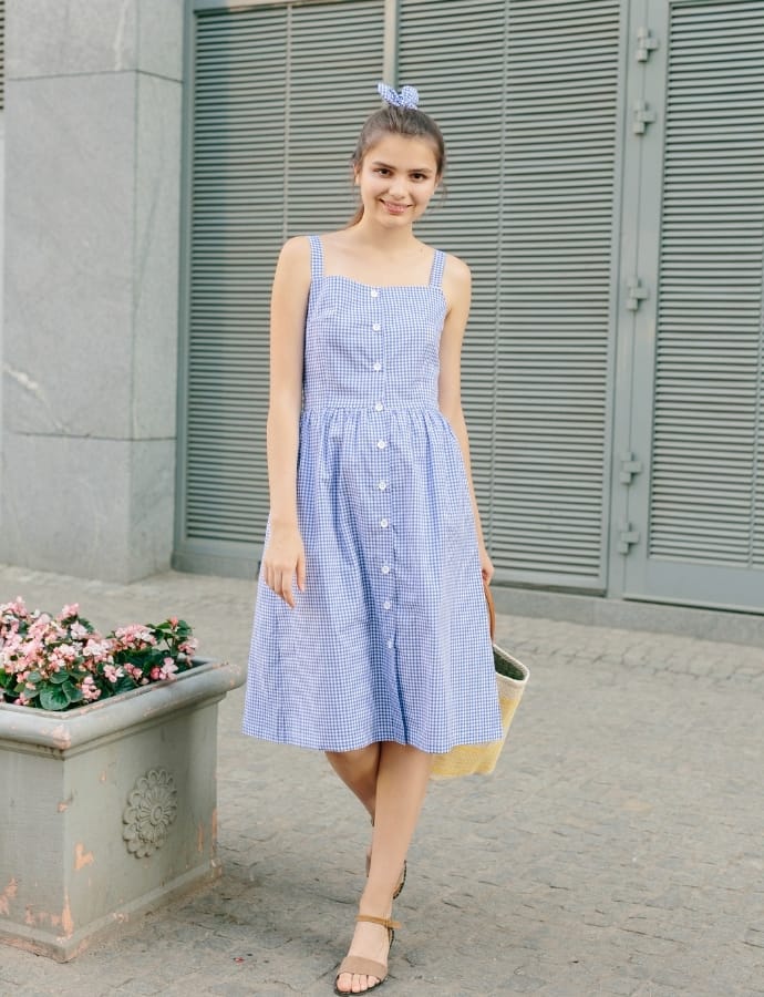 Young woman wearing a blue and white checkered cotton babydoll dress with spaghetti straps, standing outdoors near a pot of pink flowers and gray metal shutters, smiling and holding a beige tote bag.