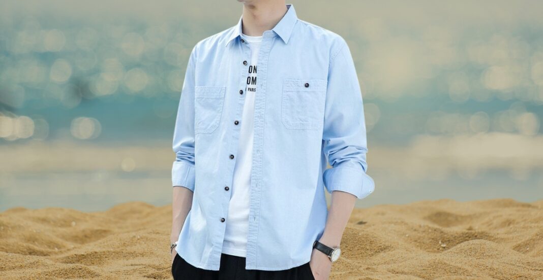 A person standing on a sandy beach, wearing a light blue cotton button-down shirt over a white t-shirt, with rolled-up sleeves.