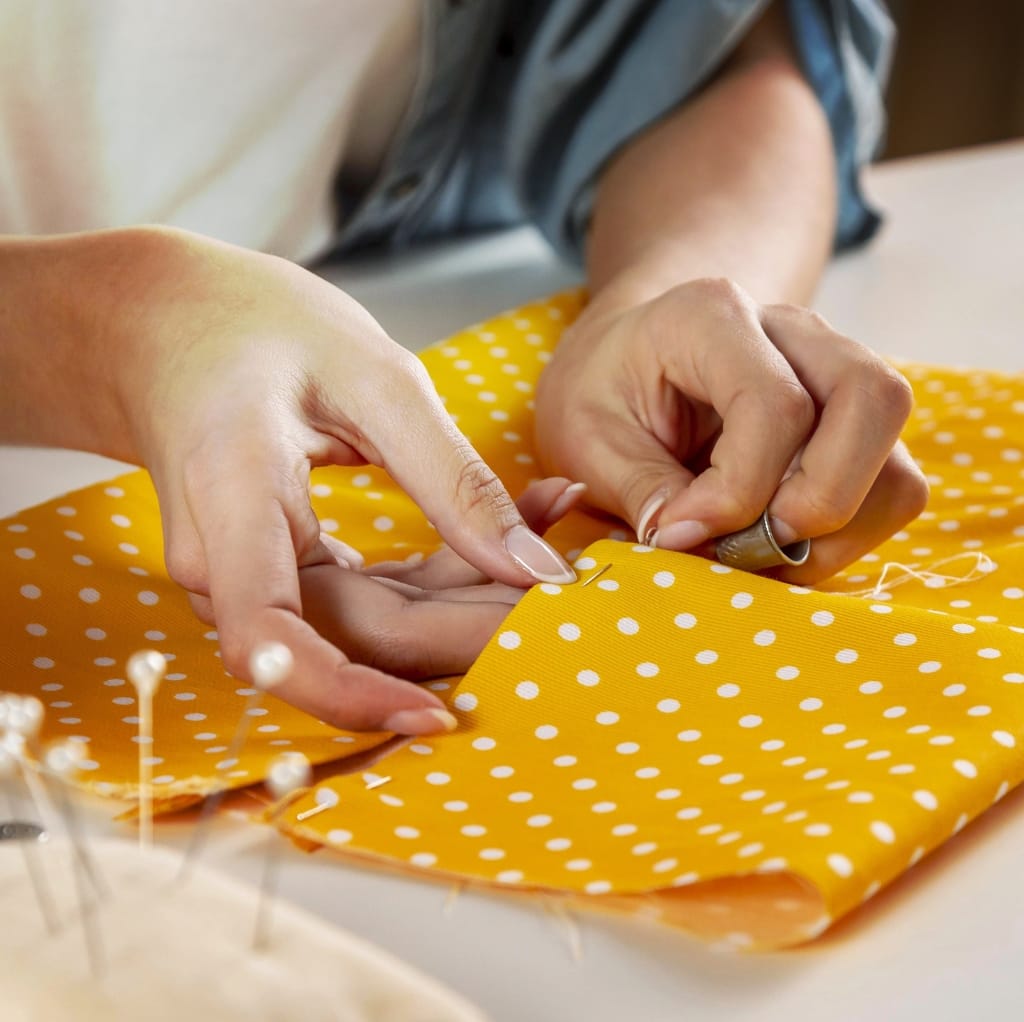 Close-up of hands sewing yellow fabric with white polka dots, using a needle and thread, likely for a first-time sewing project.