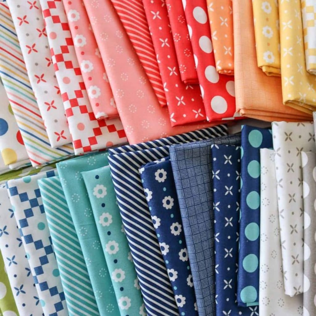 A colorful variety of cotton fabric prints arranged in rows, featuring floral, checkered, striped, polka dot, and geometric patterns in shades of red, pink, orange, yellow, blue, and white.