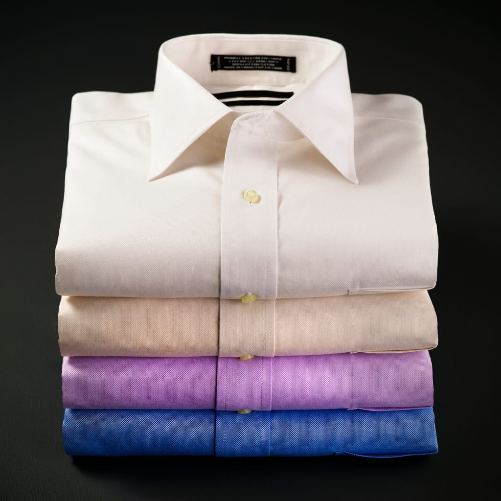 A stack of four neatly folded cotton slub shirts in white, beige, purple, and blue colors against a dark background.