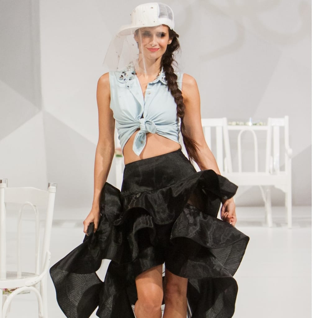 A woman walking confidently on the runway in a fashion show, wearing a sleeveless knotted top and a voluminous black skirt, with a clear face shield and cap, demonstrating the use of Crystal Organza fabric in the Catwalk Secret collection.