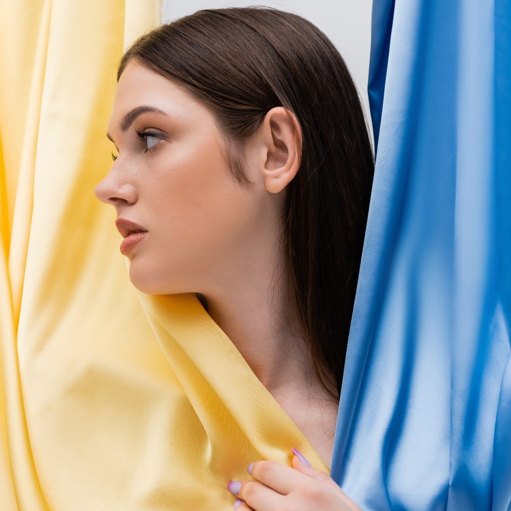 Close-up of a woman with brown hair and light skin, holding a piece of yellow fabric near her neck, standing next to blue and yellow curtains.