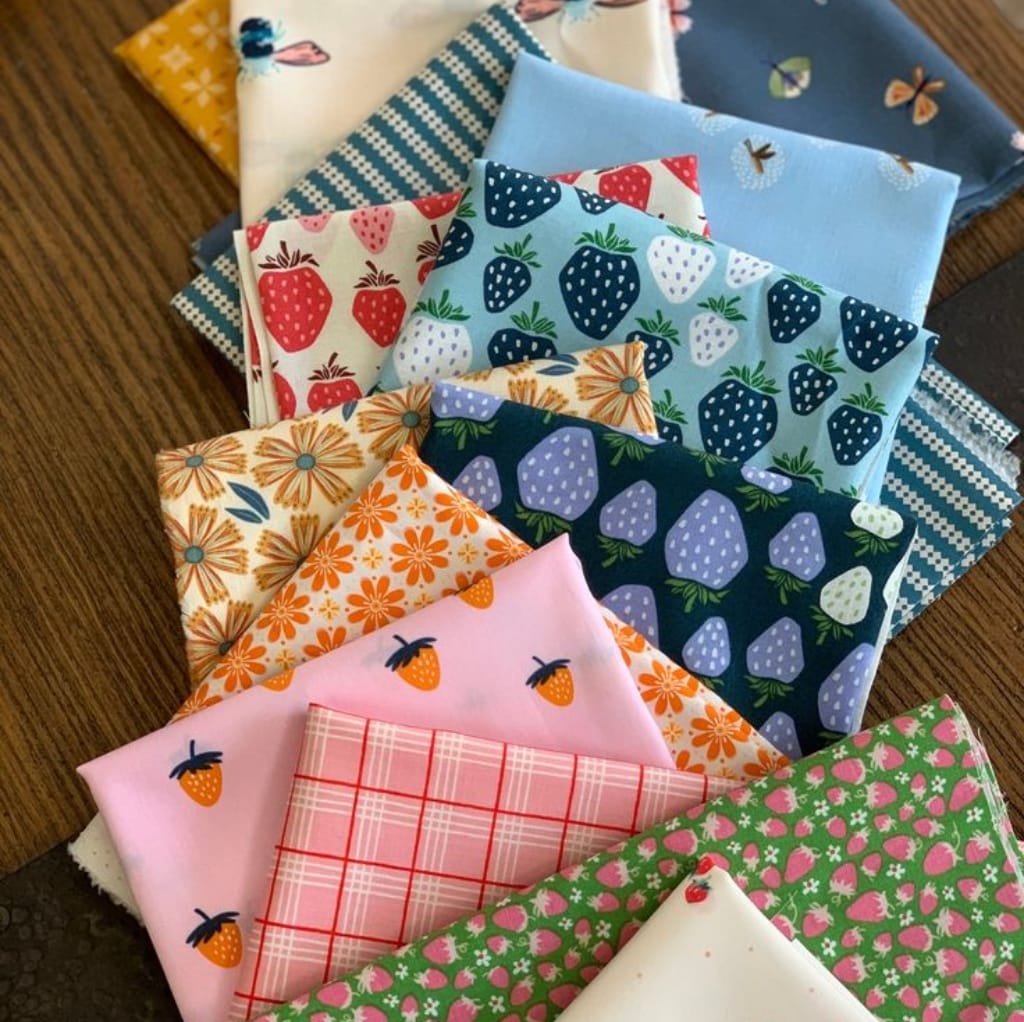A colorful assortment of cotton fabric pieces with various playful, vibrant patterns featuring strawberries, strawberries and flowers, daisies, plaid, strawberries with a pink background, and tiny pink motifs on a green background.