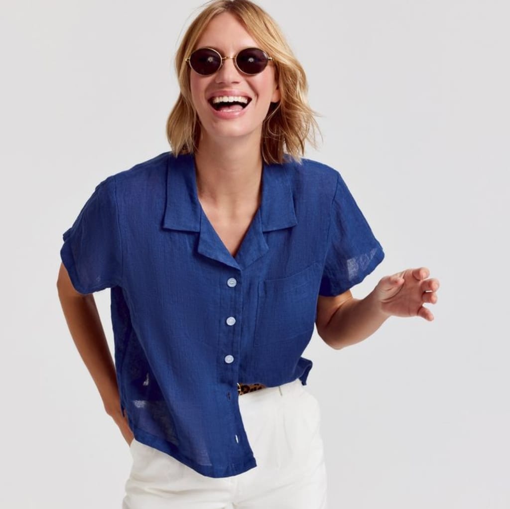 A woman wearing a blue short-sleeve linen shirt, smiling and posing against a plain background.