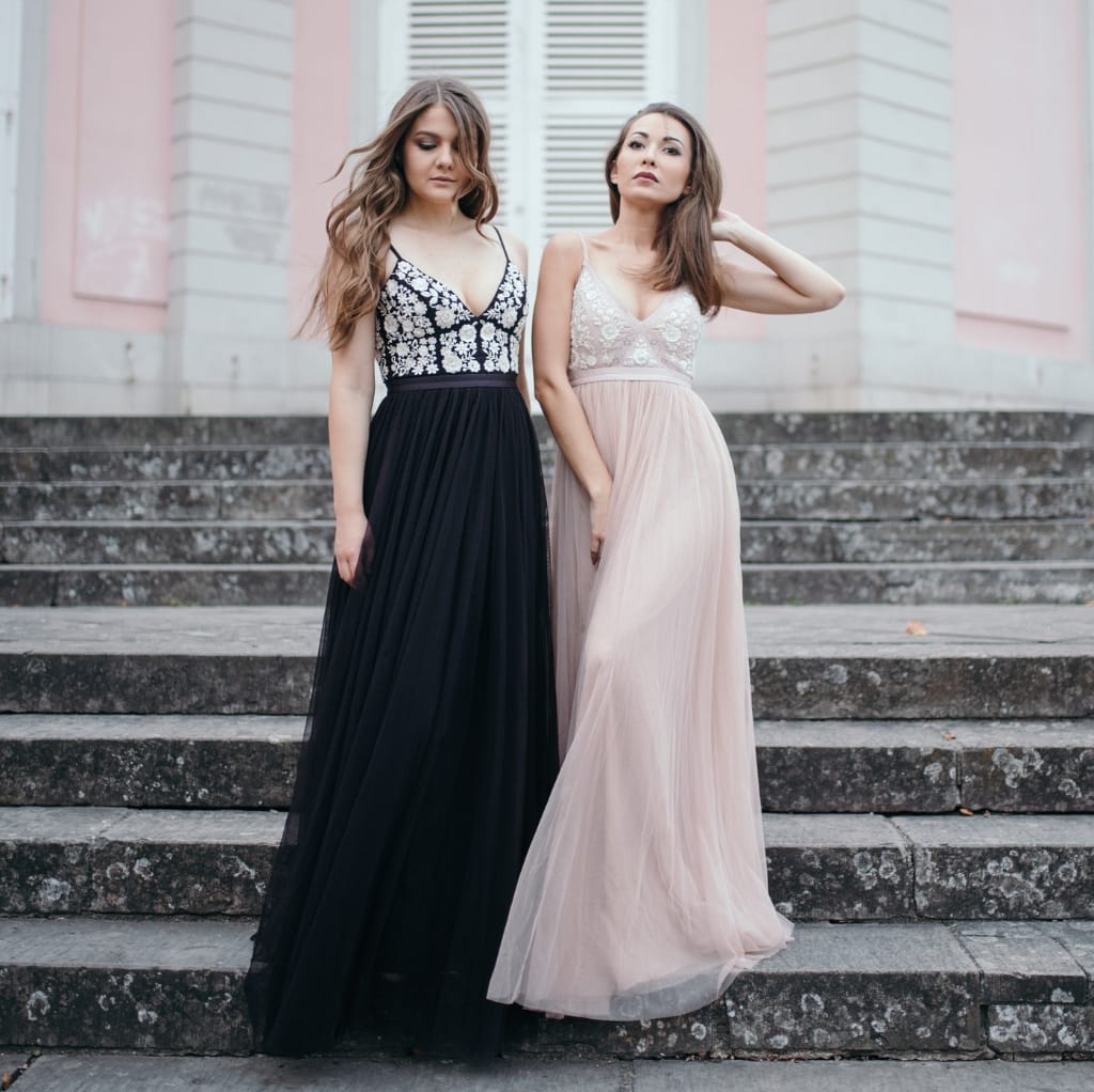 Two women standing on stone steps outside a building, wearing elegant, flowing evening dresses. The woman on the left is in a black dress with a lace, floral-patterned bodice, while the woman on the right is in a light pink, chiffon dress with a delicate, embroidered bodice.