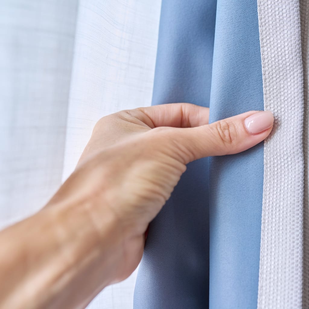 Close-up of a hand gently touching thick, light blue, and textured cotton fabric hanging vertically among other fabric swatches.