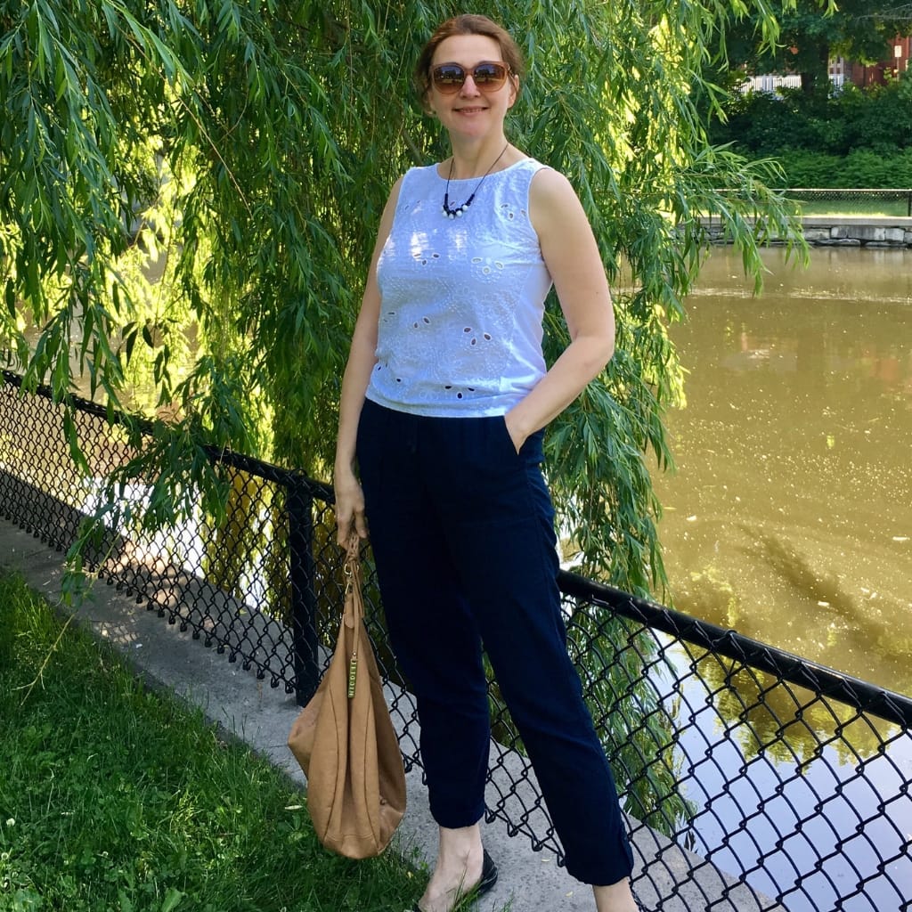 A woman stands outdoors near a pond, wearing a casual chambray blouse and dark chambray trousers. She has sunglasses, a beaded necklace, and carries a tan bag. She is smiling with one hand in her pocket, surrounded by lush greenery and sunlight.