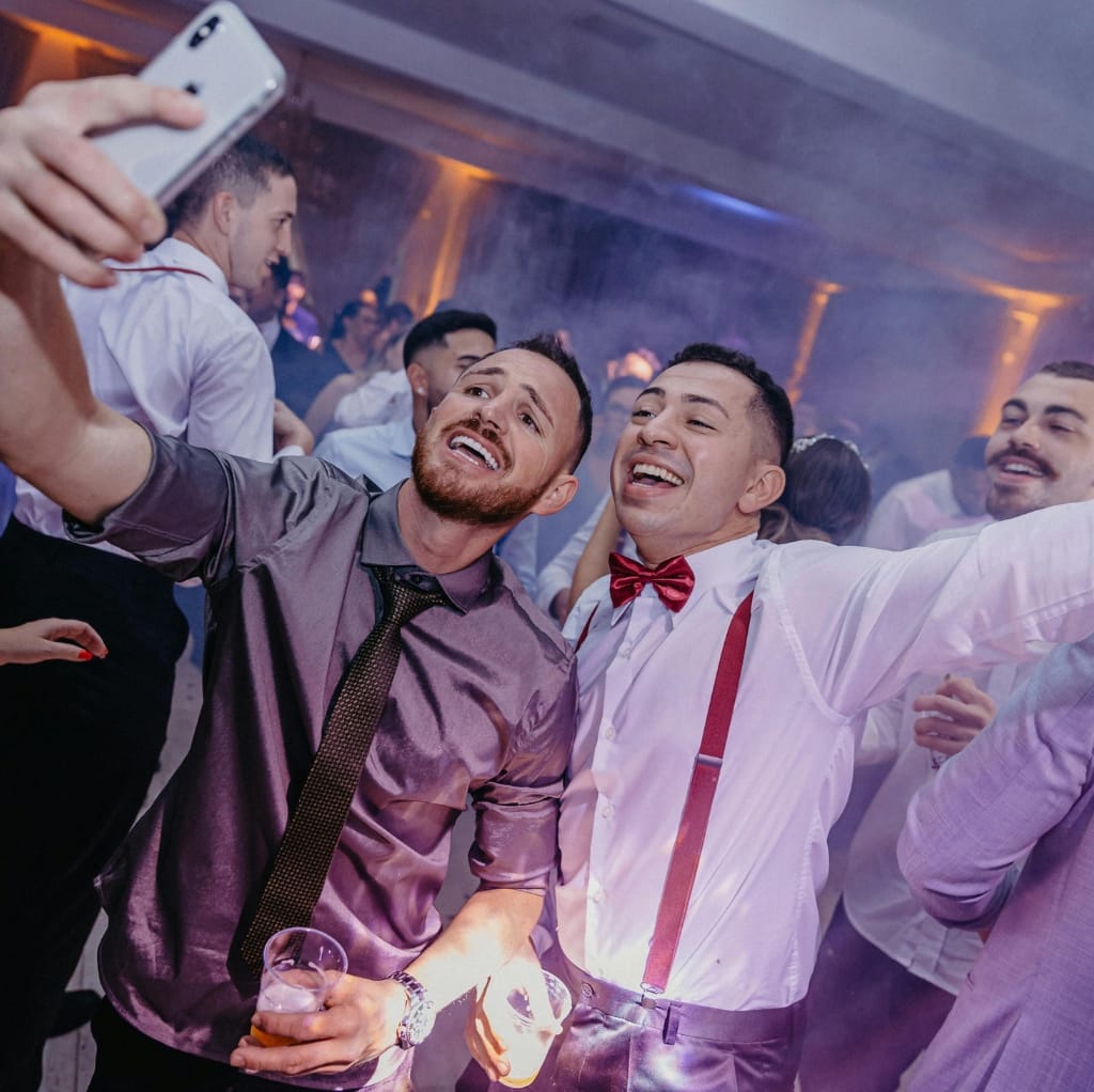 Two men taking a selfie at a lively evening party, dressed in casual linen shirts. One wears a shiny, dark purple linen shirt with a black tie, holding a drink, while the other wears a white linen shirt with red suspenders and a red bow tie, smiling and posing for the photo. Other guests are seen dancing and enjoying the event in the background.
