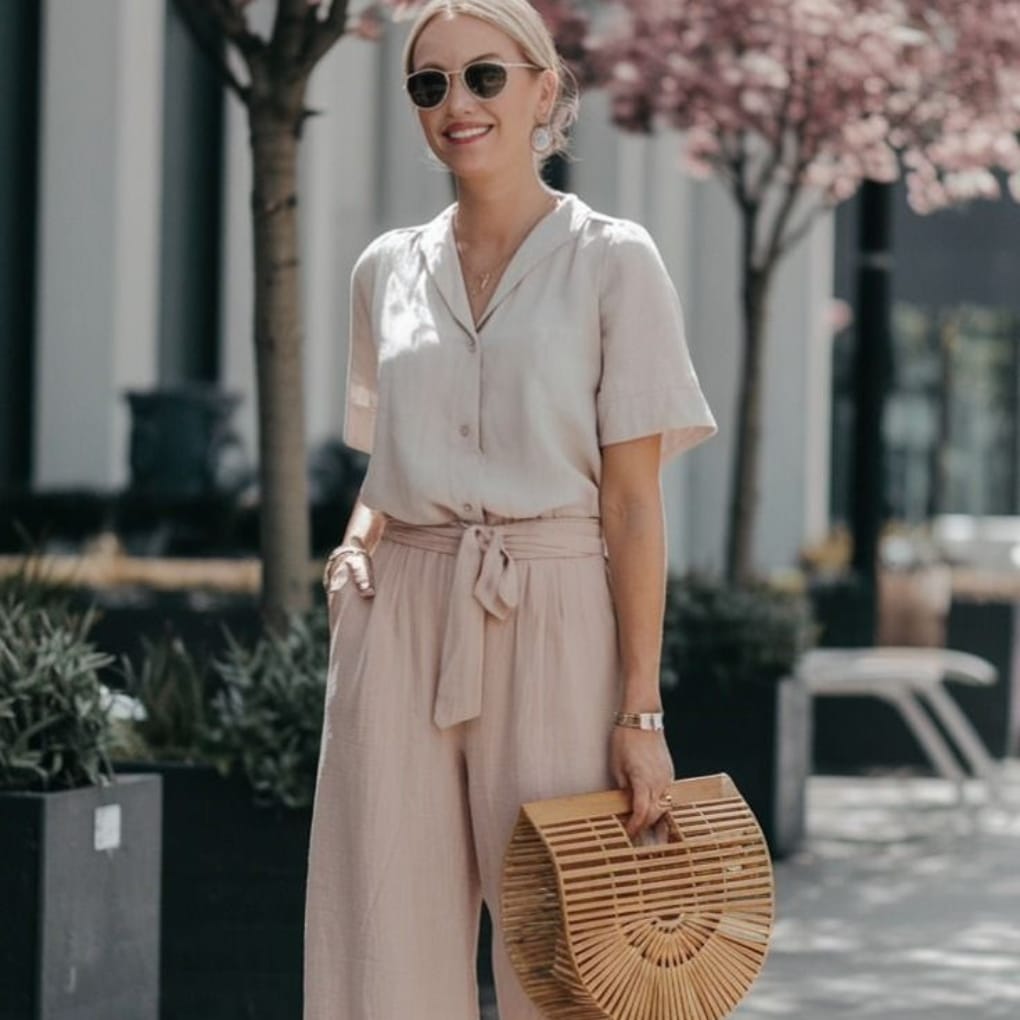 Woman with blonde hair wearing sunglasses, a light beige short-sleeve blouse, matching wide-leg pants, and holding a wooden bamboo handbag, walking outdoors with pink flowering trees in the background.